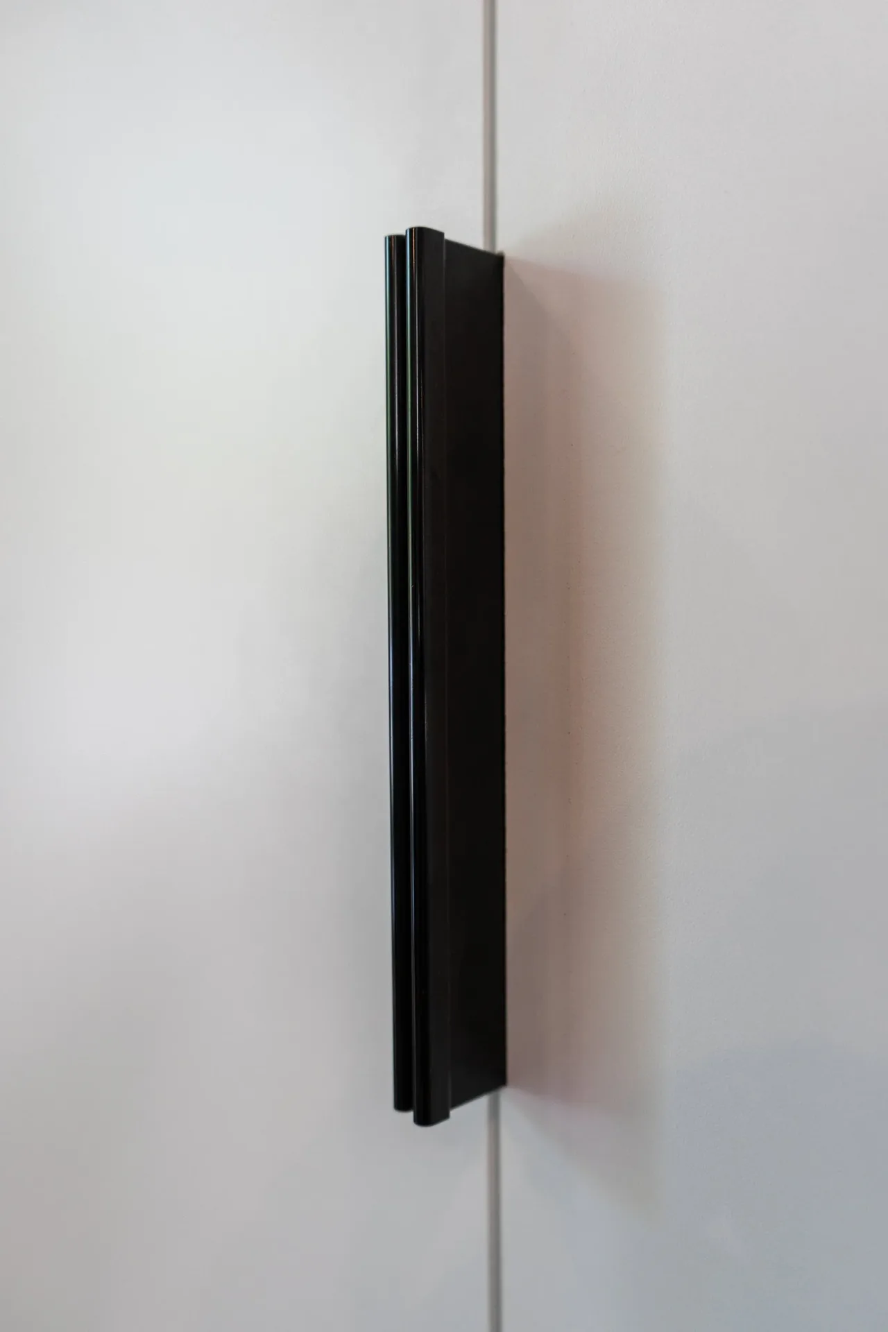 A black vertical handle is mounted on a smooth white cabinet door.