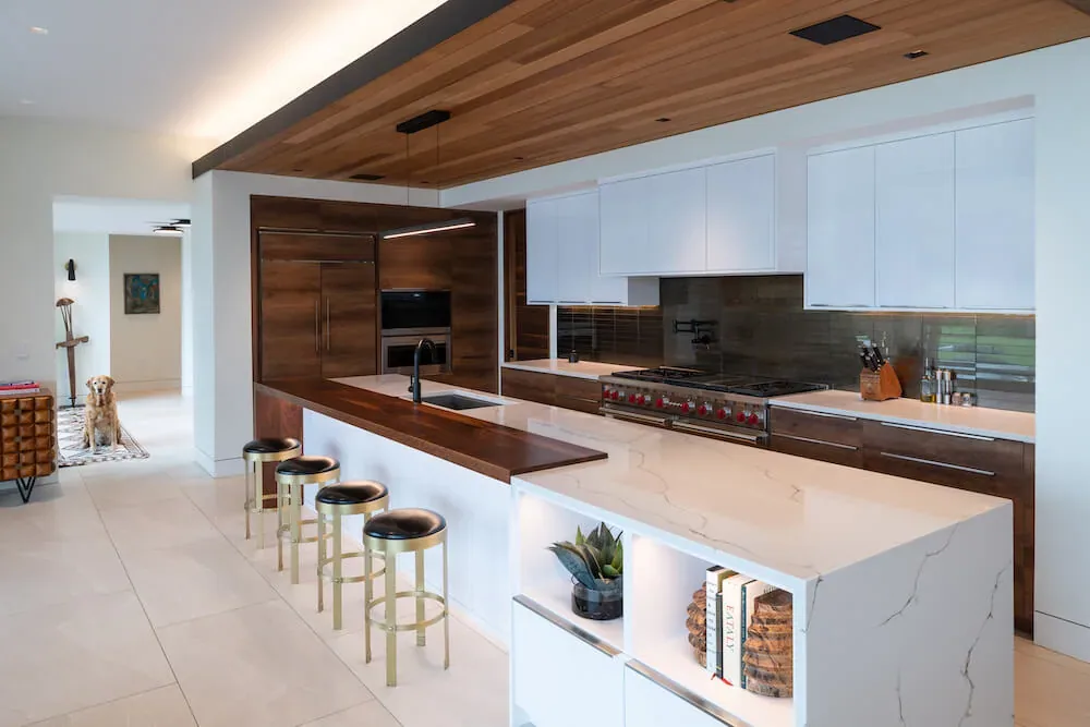 The Pelican House kitchen features a marble island, wood and white cabinetry, and a wood-paneled ceiling accent with a linear pendant light.