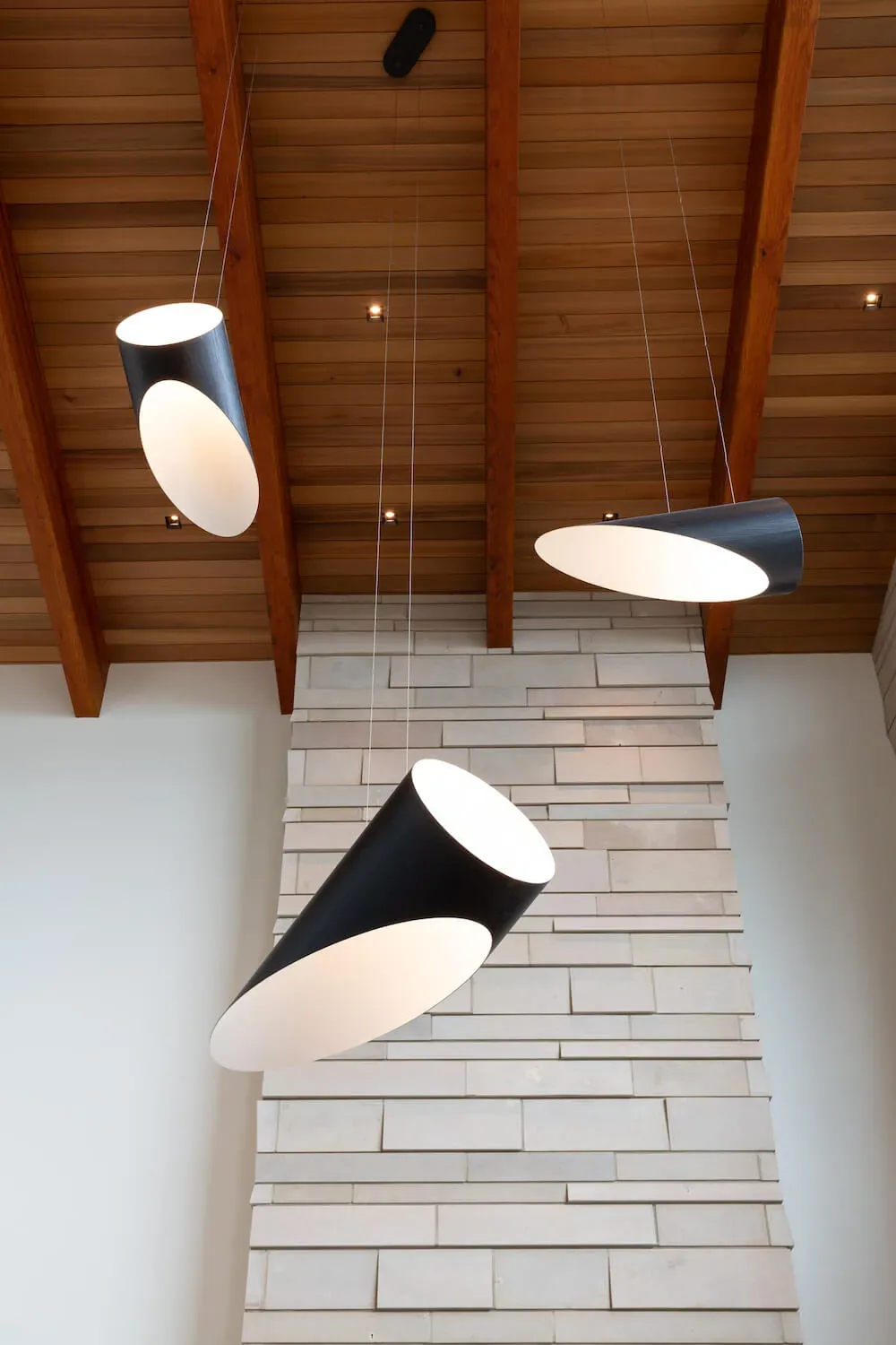 Modern pendant lights hang from a wood-paneled ceiling with exposed beams above a stacked stone wall.