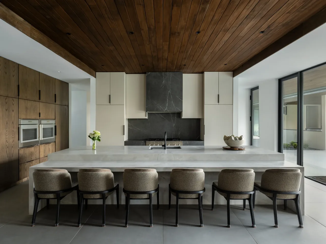 This kitchen features a white island with seating for six, a wood-paneled ceiling, and black framed glass doors opening to an outdoor patio.