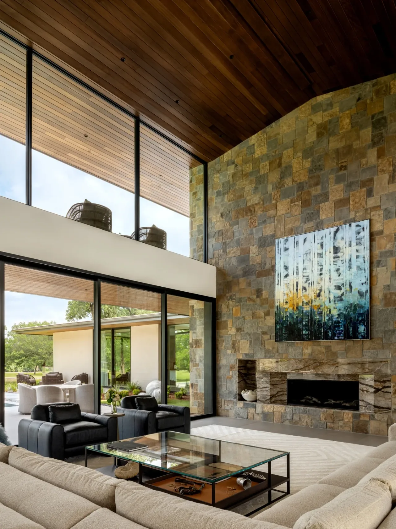 The living room features a tall stone fireplace with a marble hearth, a beige sofa, black leather armchairs, and floor-to-ceiling glazing.