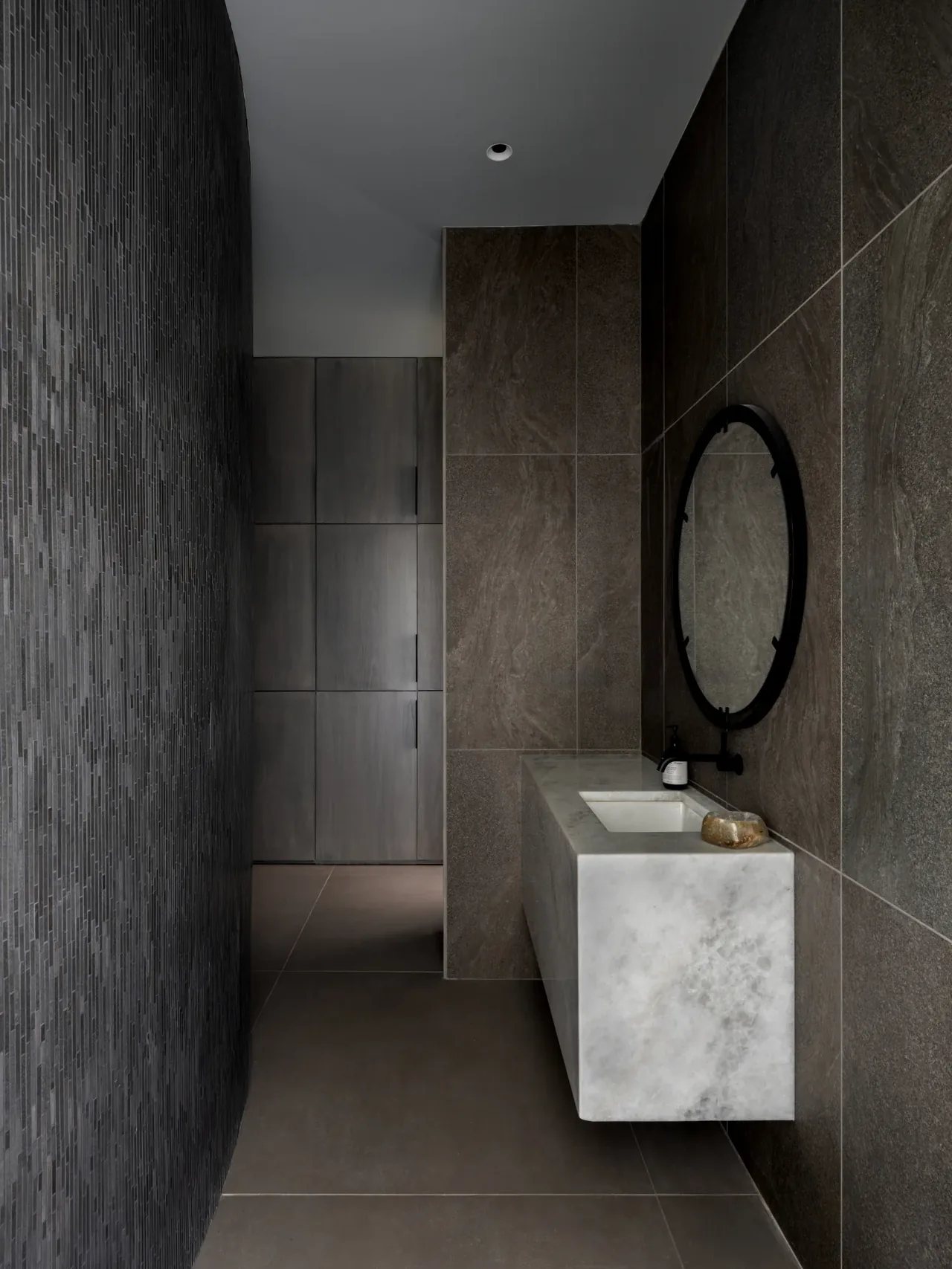 Bathroom featuring dark tile walls, a floating marble vanity, and a round black-framed mirror.