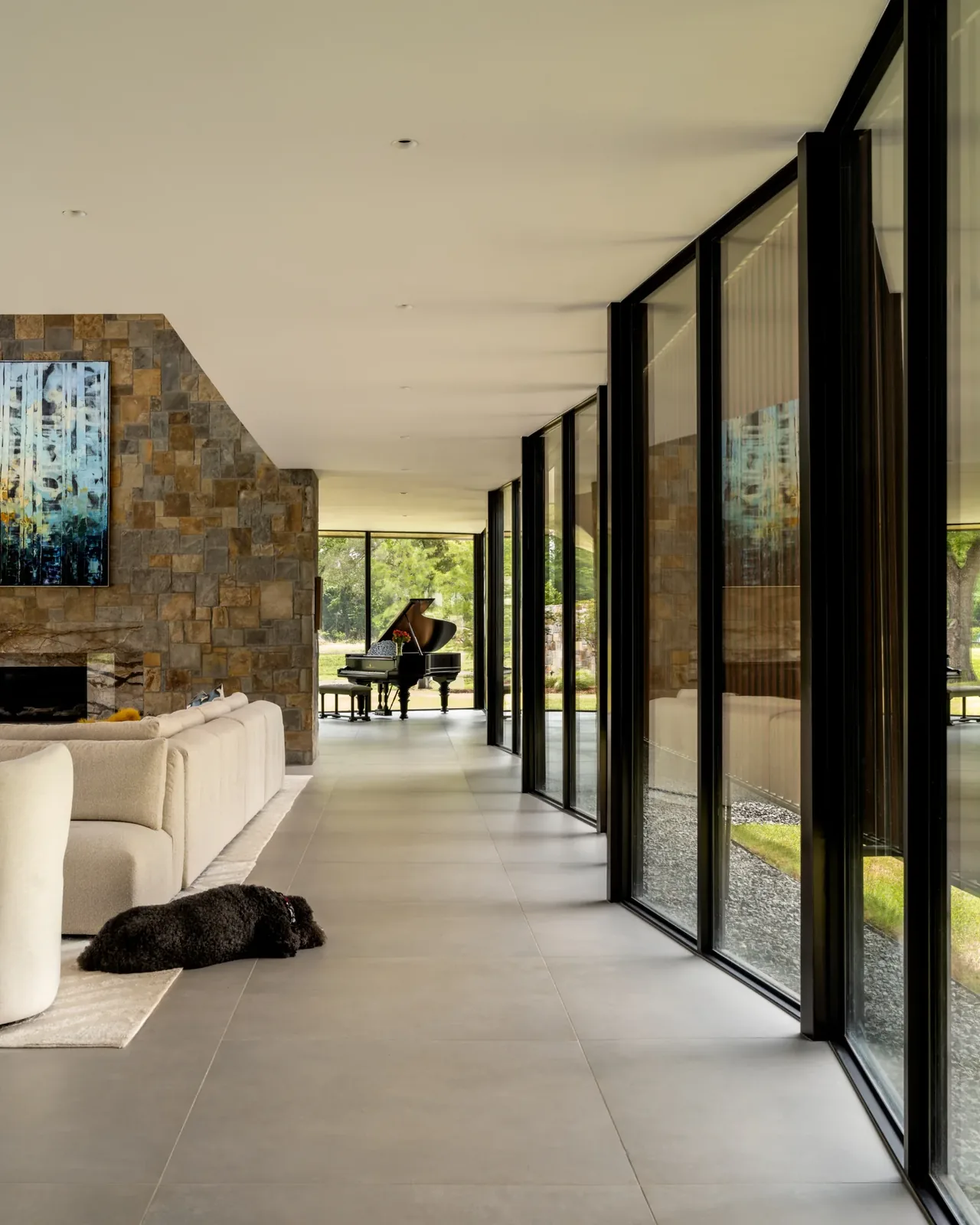 Interior corridor with floor-to-ceiling windows, stone fireplace wall, beige seating, and a grand piano at the end.