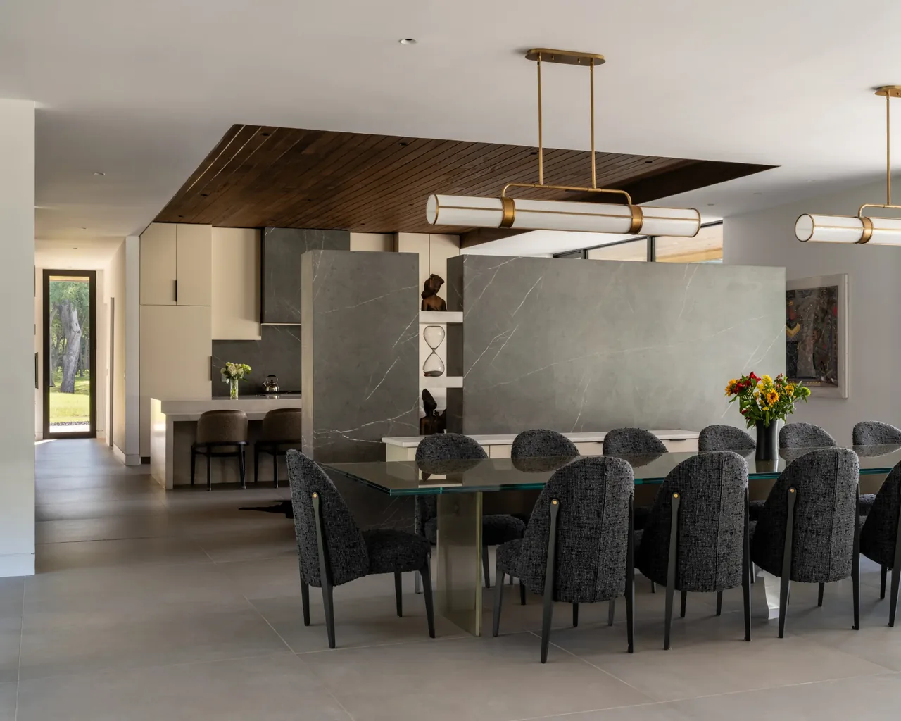 Dining room with a glass table and gray upholstered chairs, separated from the kitchen by a stone wall with display niches.