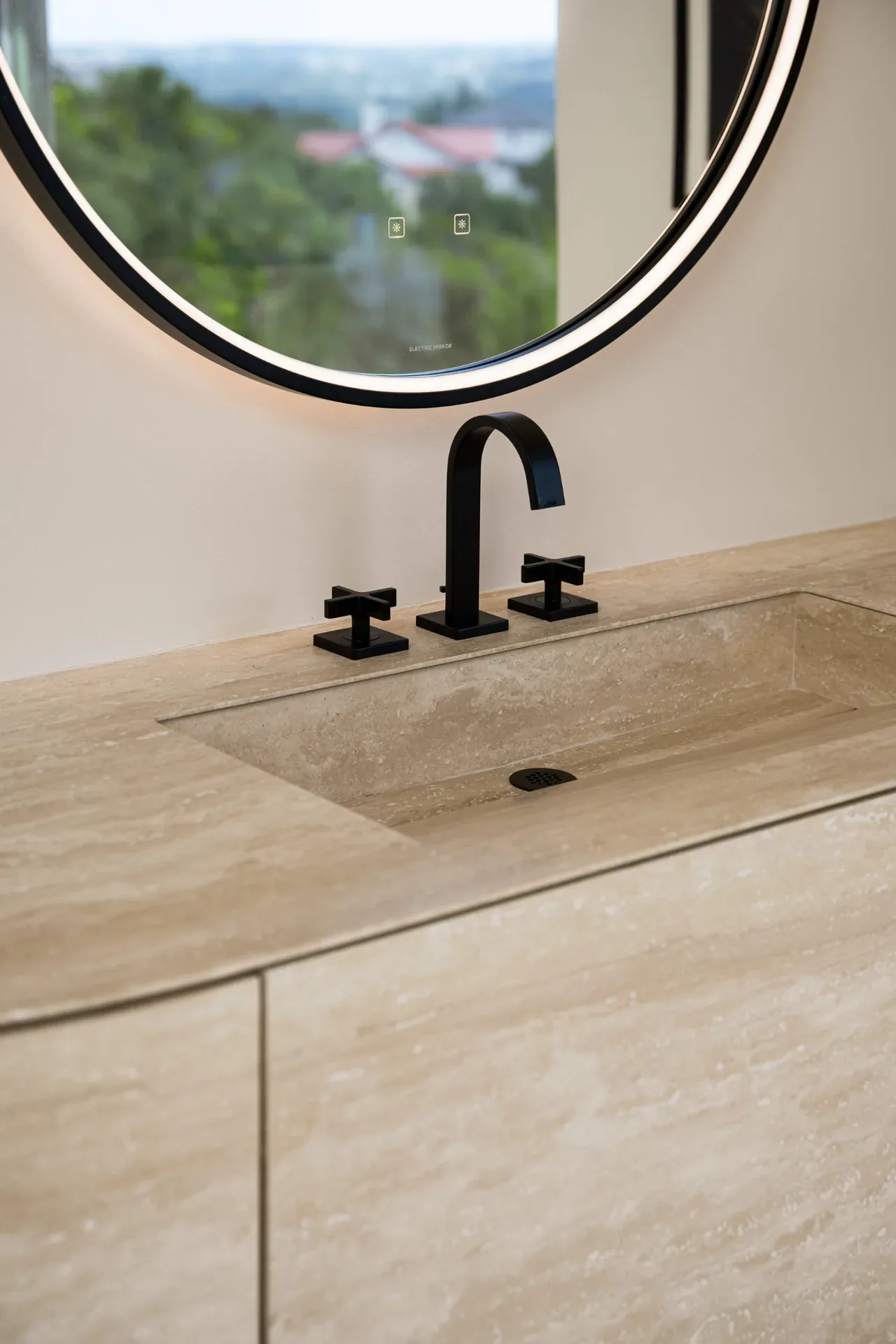 A bathroom sink with a beige stone countertop, matte black faucet, and round illuminated mirror is shown in the Villa Morada project.