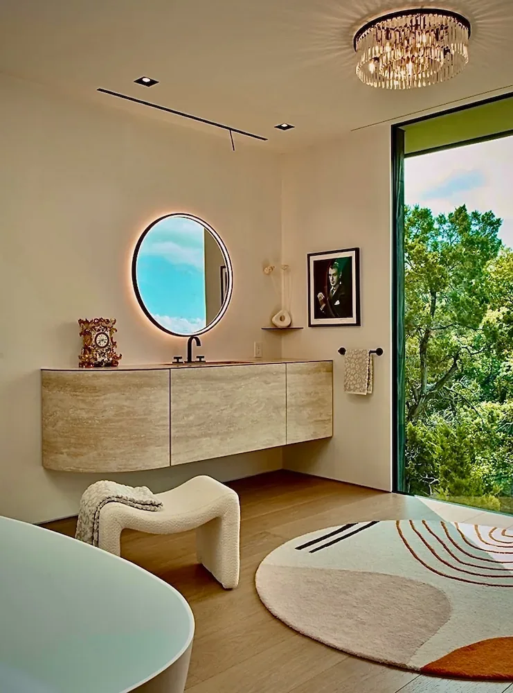 Villa Morada bathroom by Winn Wittman Architecture features a floating wood vanity and round backlit mirror with a view of lush trees outside.