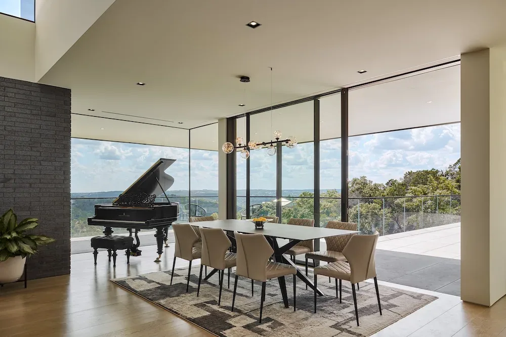 Villa Morada dining area featuring a large table, chandelier lighting, patterned rug, and floor-to-ceiling glazing overlooking a hill landscape.