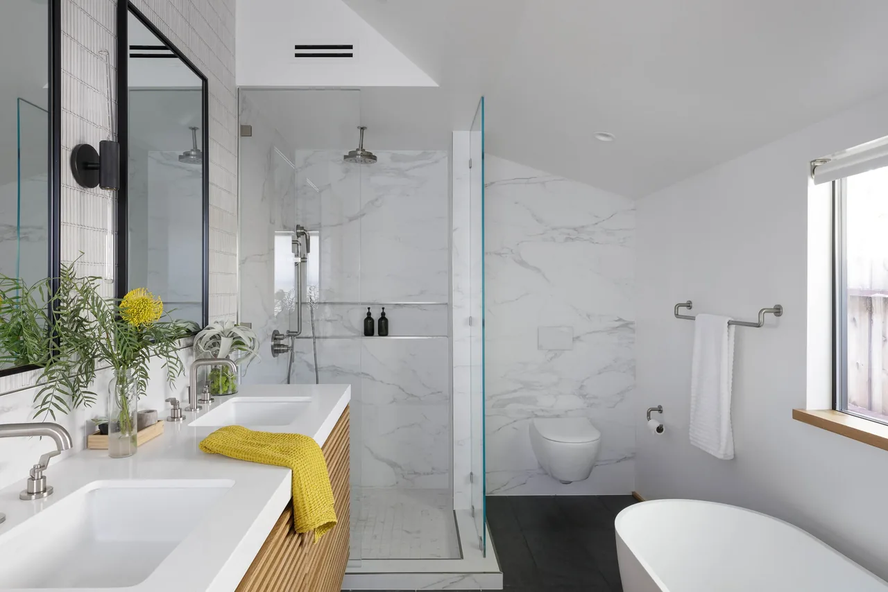 The bathroom features dual sinks with wood cabinetry, marble walls in the shower, and a standalone bathtub near a window.