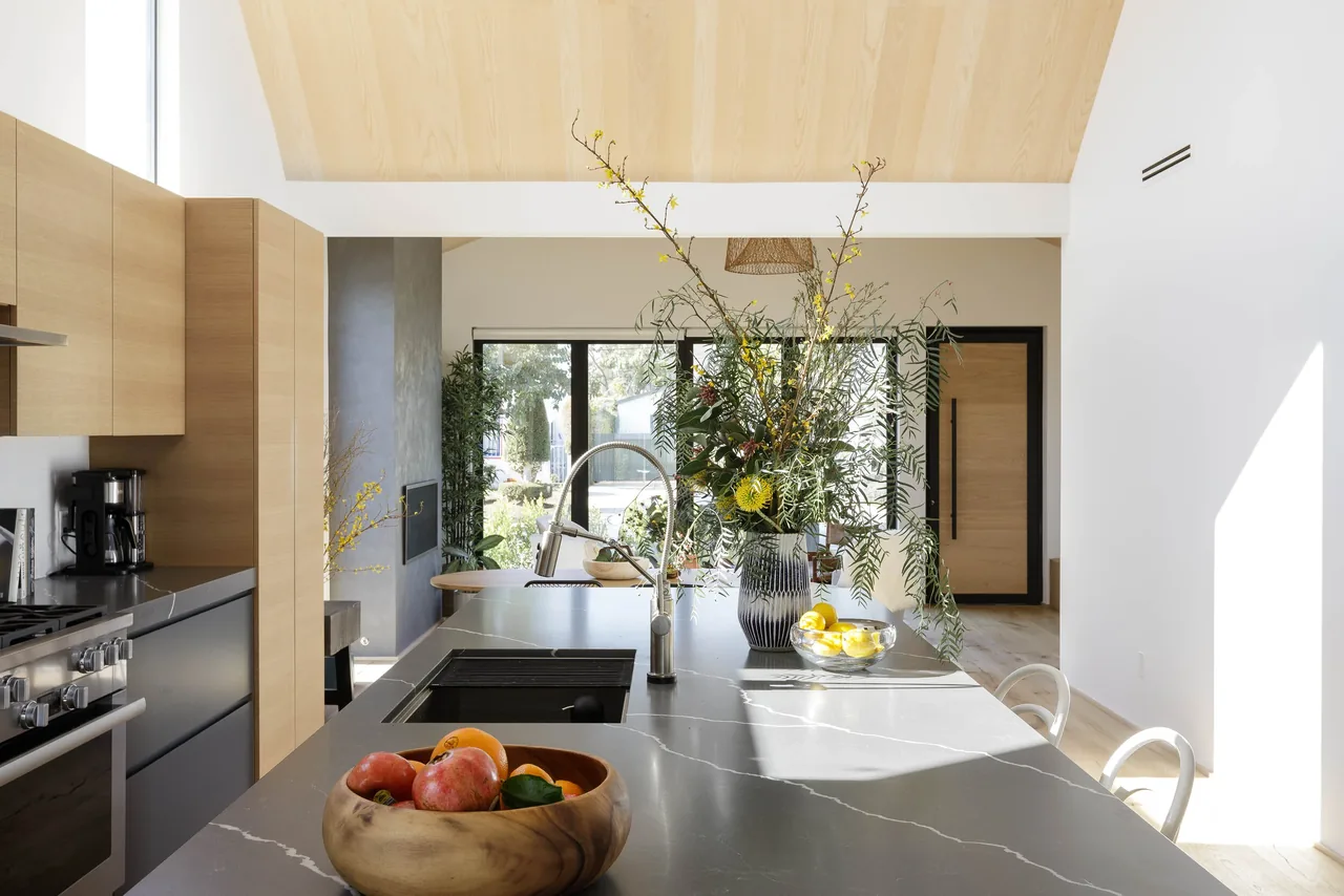 Curson Residence kitchen features a vaulted wood paneled ceiling, large island with sink, and black window frames around sliding glass doors.