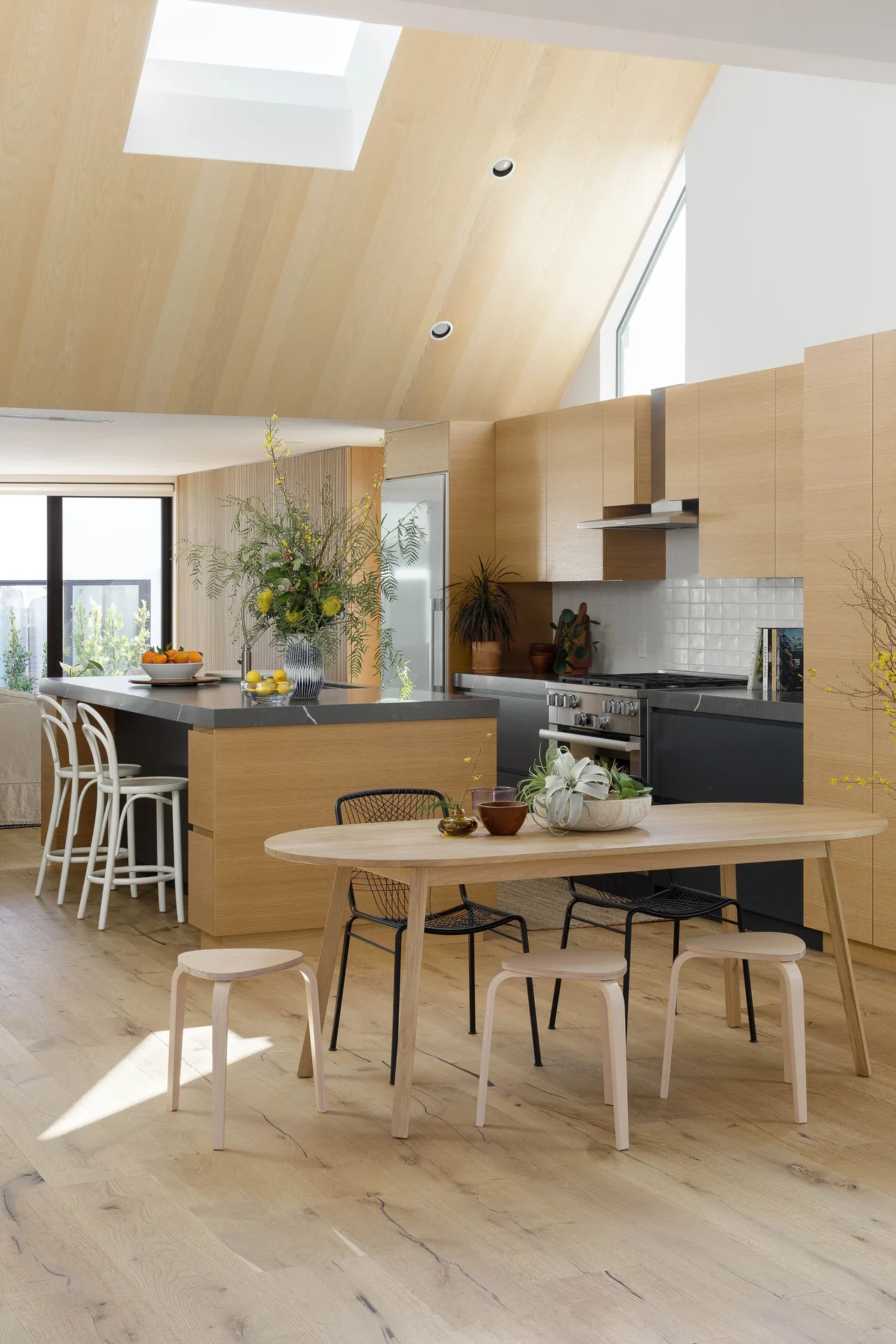 The Curson Residence kitchen and dining area feature wood cabinetry, a skylight, and a mix of black and light wood chairs.