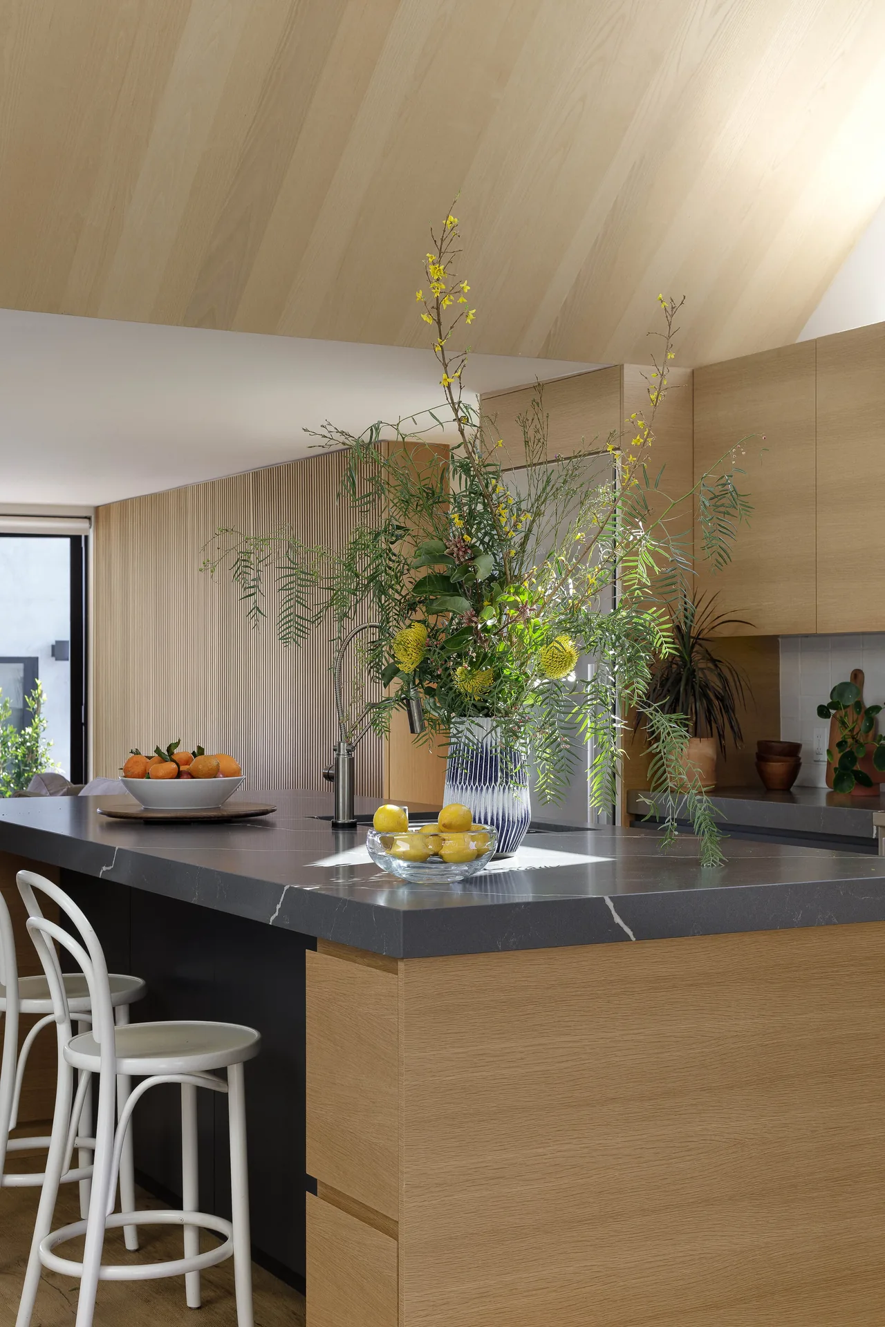 Curson Residence kitchen features wood cabinetry and a large dark stone countertop island with bar stools, decorated with fresh flowers and fruit.