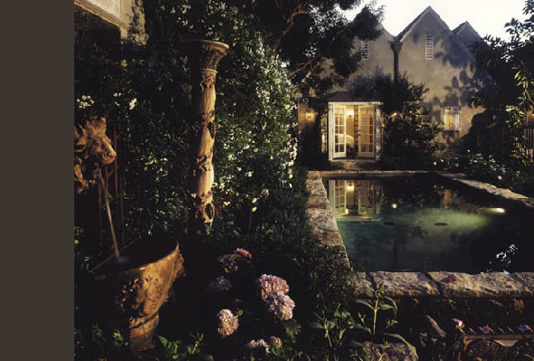 Stone-bordered pool and garden with plants outside a lit home at night.