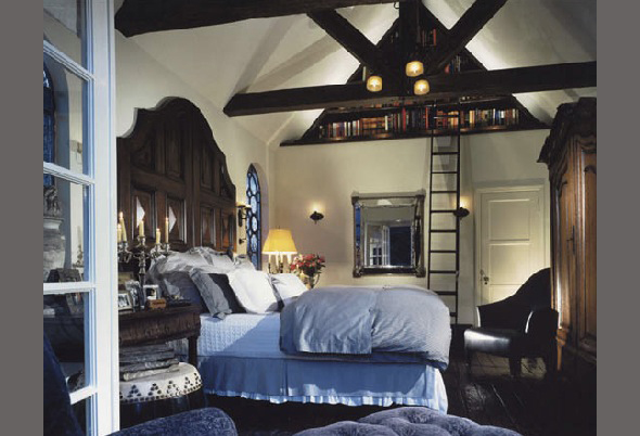 Bedroom featuring vaulted ceiling with exposed beams, blue bedding, vintage-style wooden furniture, and a study ladder to a high bookshelf.