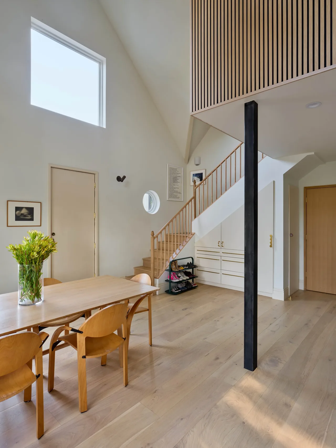 Dining space features light wood furniture, high ceilings, a large square window, and a staircase with vertical wood slats.