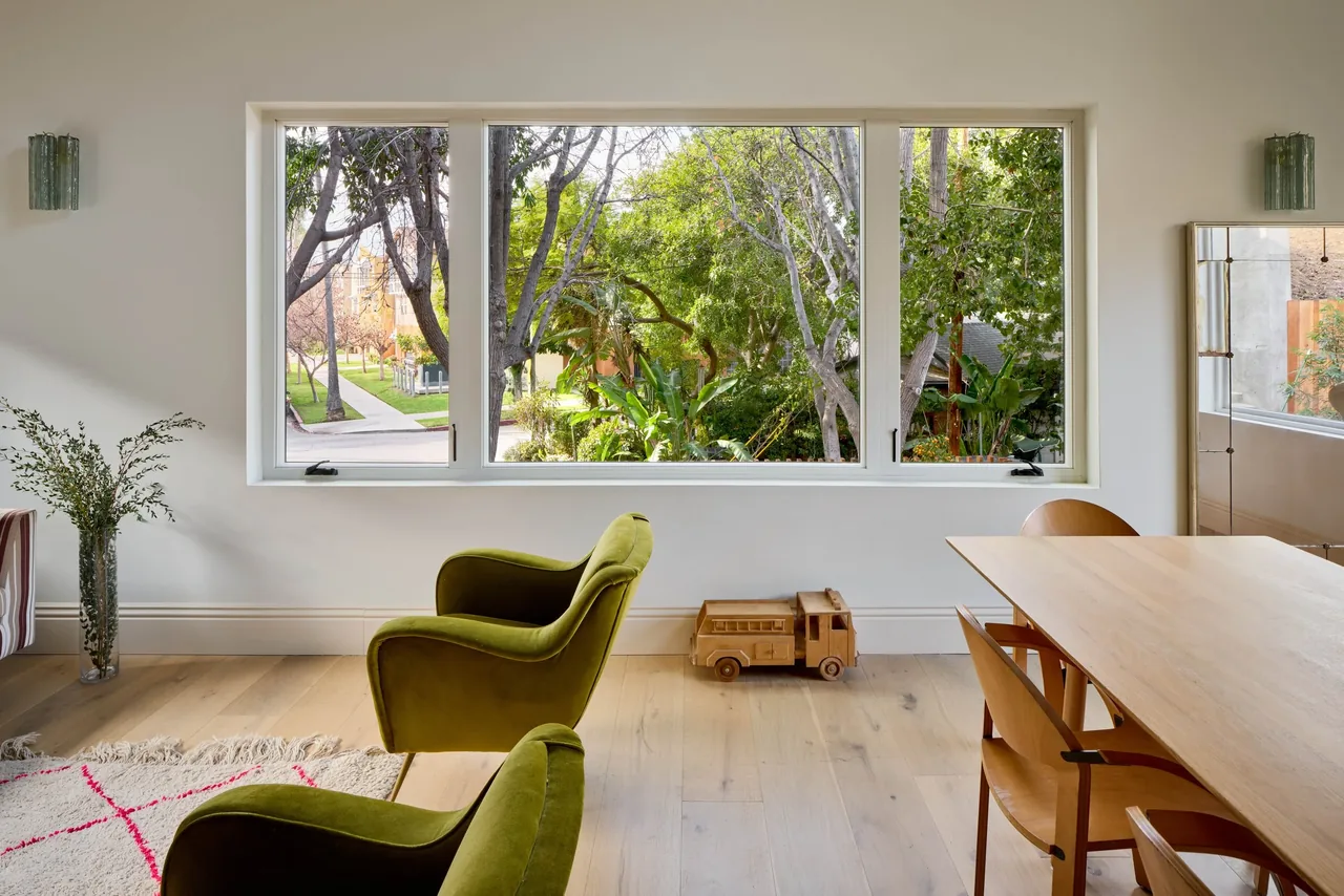 The Silver Lake Treehouse features a dining area with wooden chairs and a table, green armchairs, and a rectangular window with views of garden trees.