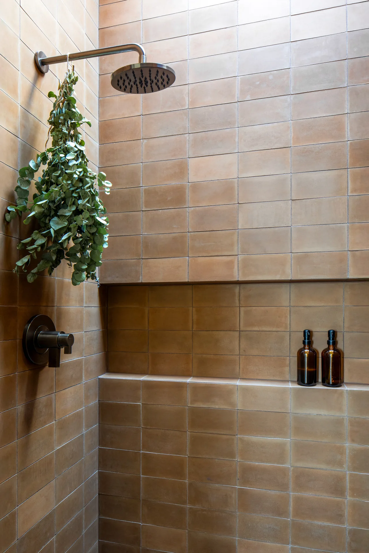 Bathroom shower area with matte brown tiles and built-in shelf holding two brown pump bottles.