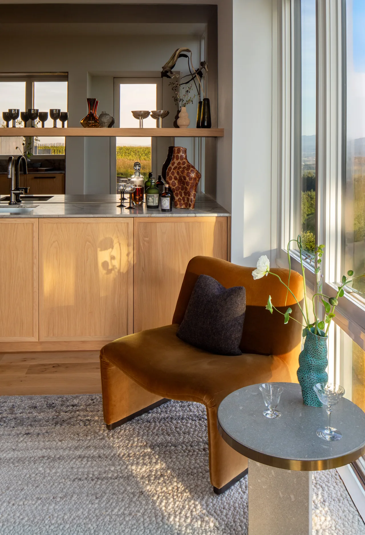 Hillside House living area with velvet chair, large wood cabinet doors, and stone countertop island, capturing the vineyard view through expansive windows.