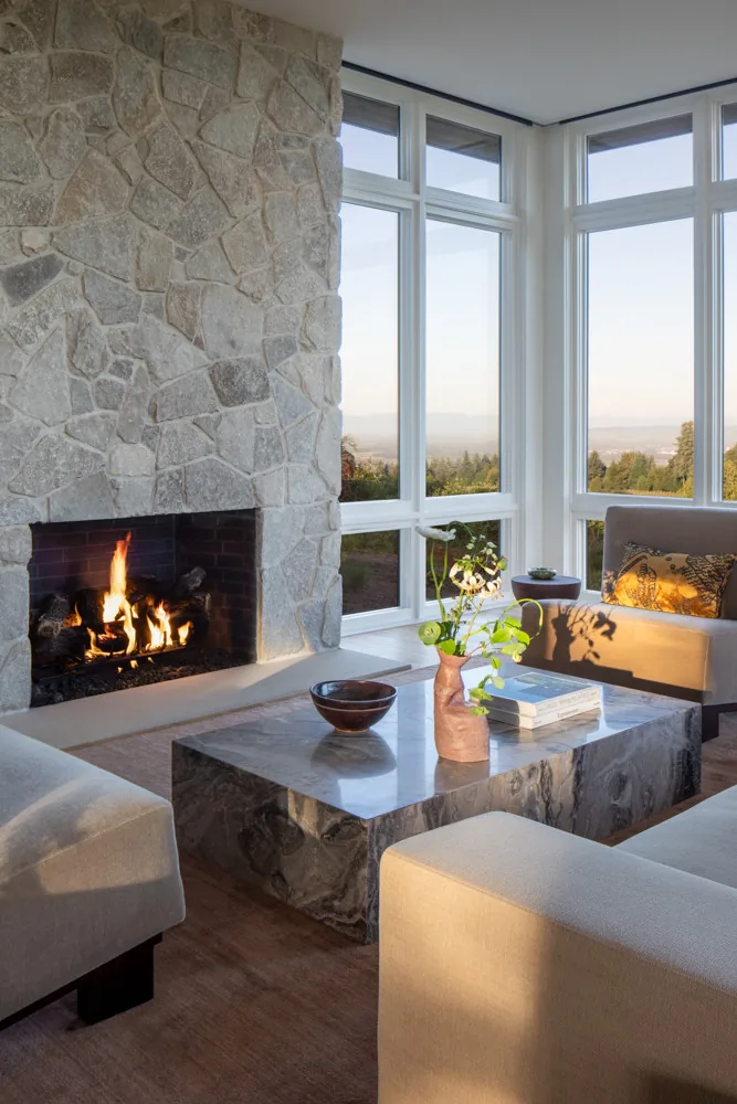 The Hillside House living room features a stone fireplace, large windows framing the landscape, and a marble coffee table.