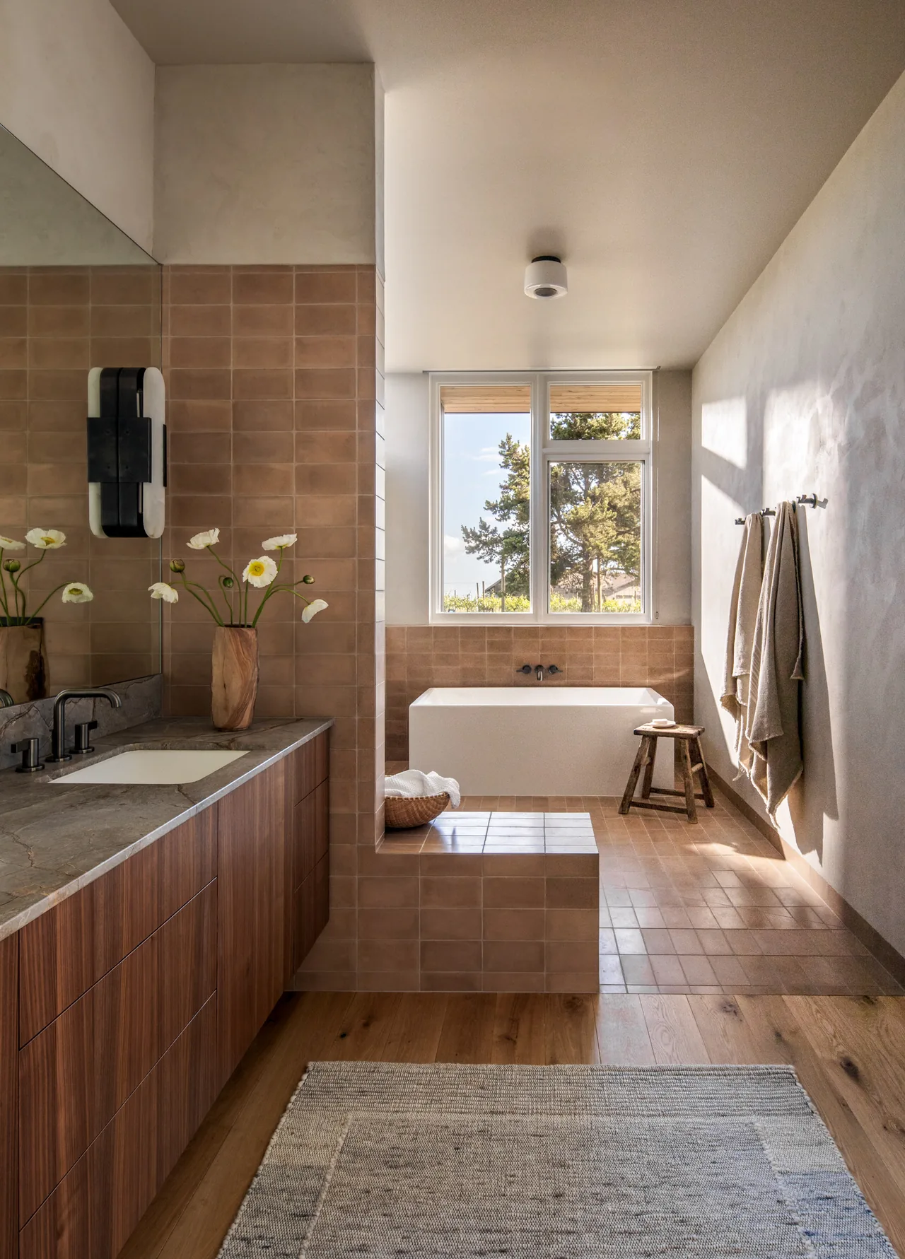 Hillside House bathroom features a built-in tub with beige tile surround and a wooden vanity with marble countertop under a large window.