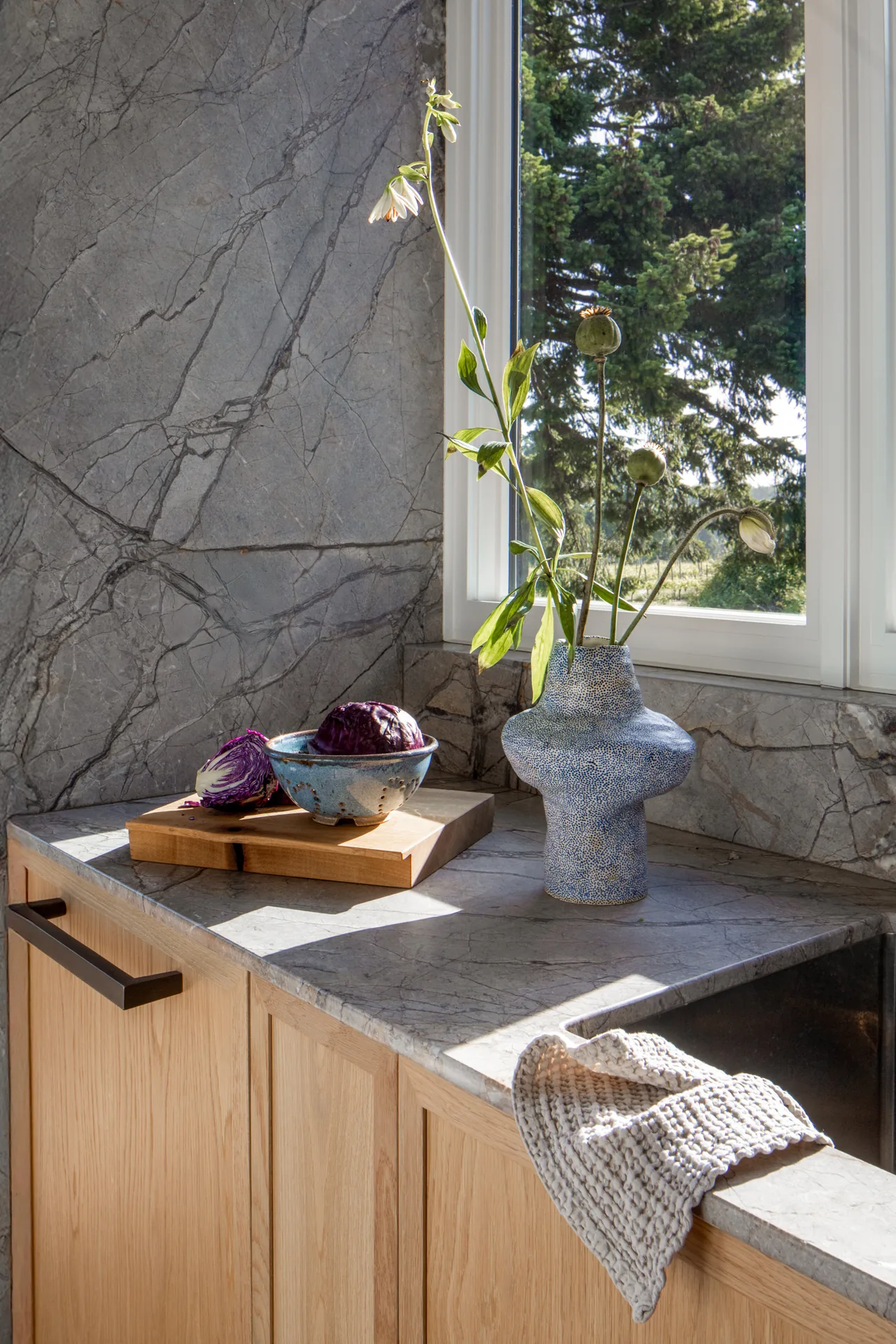 Kitchen corner featuring gray stone countertop, light wood cabinets, and a unique vase with flowers by a window.