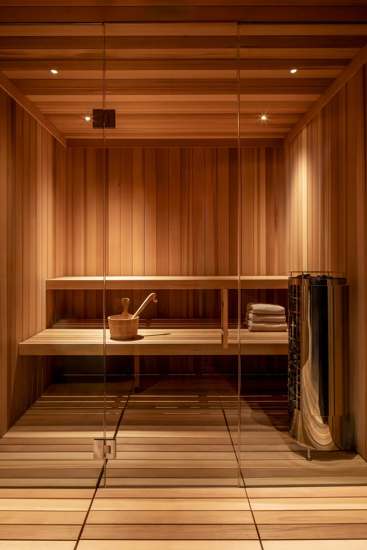Sauna room with sleek wood paneling, glass door, and metal heater featuring a wooden bucket and stacked towels.