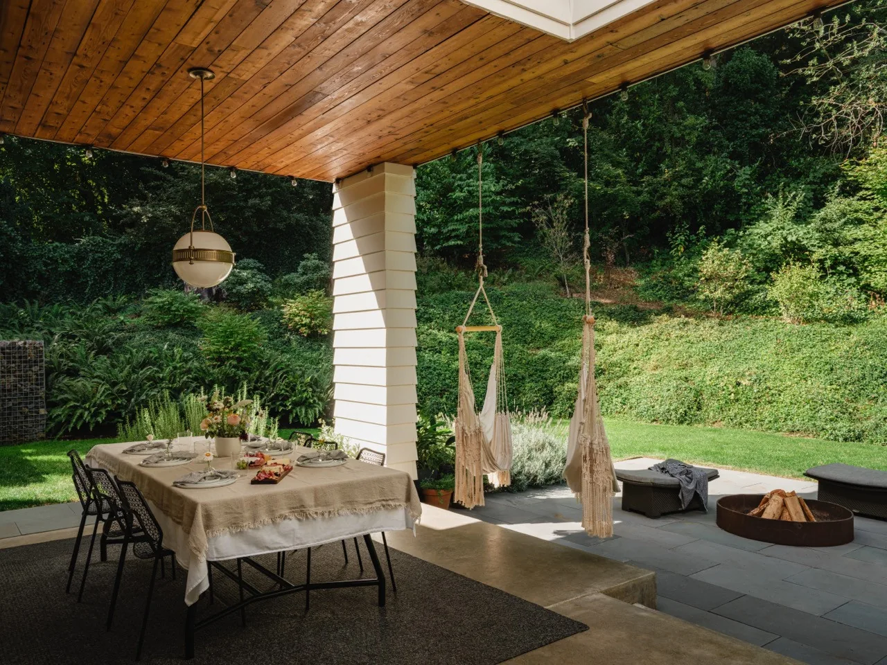 The covered patio features a wooden ceiling, hanging fabric chairs, dining table with chairs, and a fire pit overlooking lush greenery.