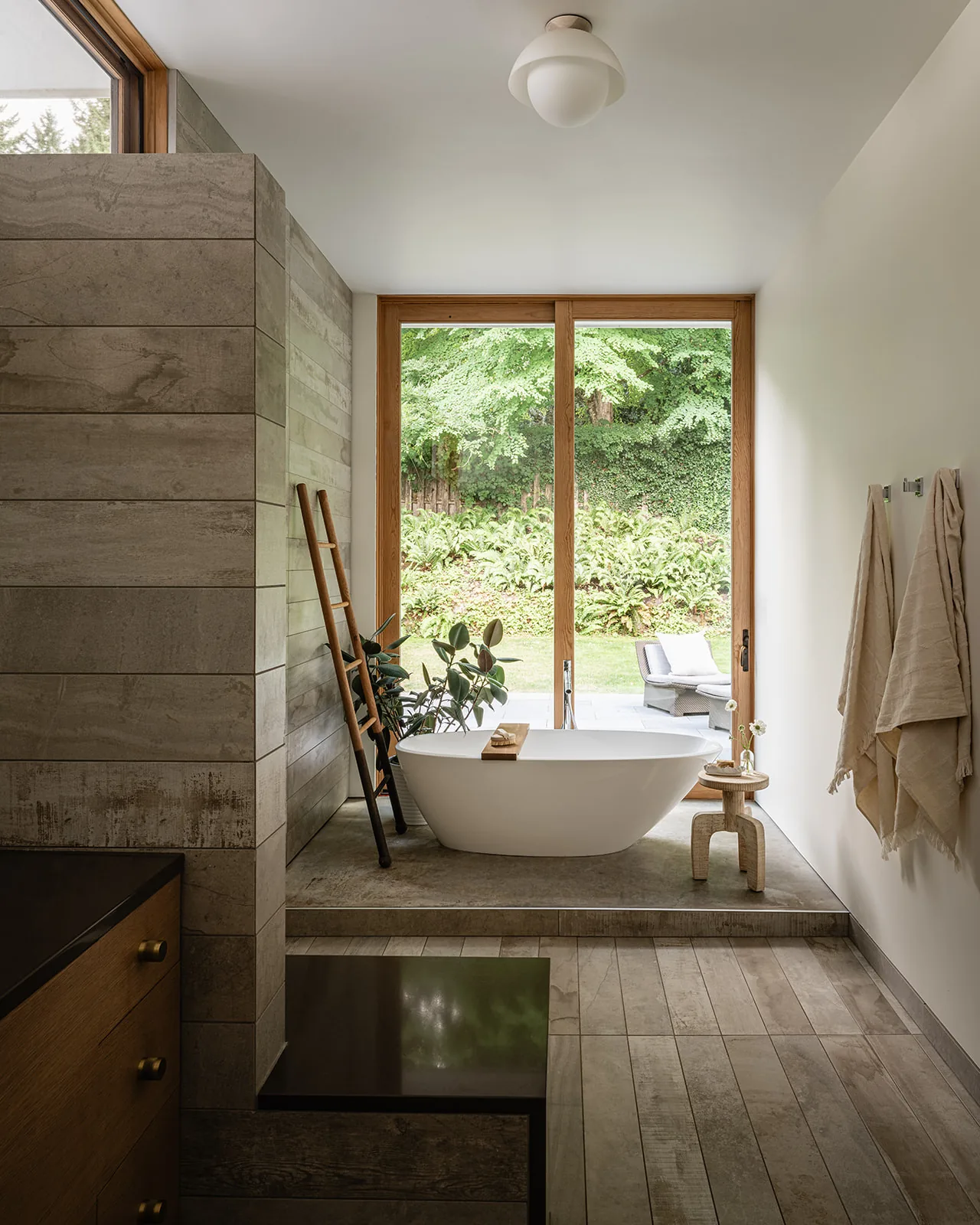 Freestanding tub in a bathroom with large wood-framed sliding glass doors facing a lush garden.
