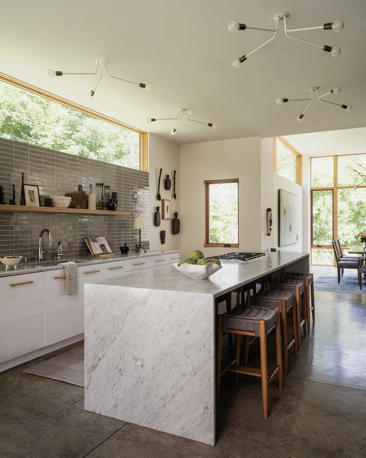 The Idea House kitchen features a white marble waterfall island, gray tiled backsplash, and natural wood window frames providing abundant daylight.