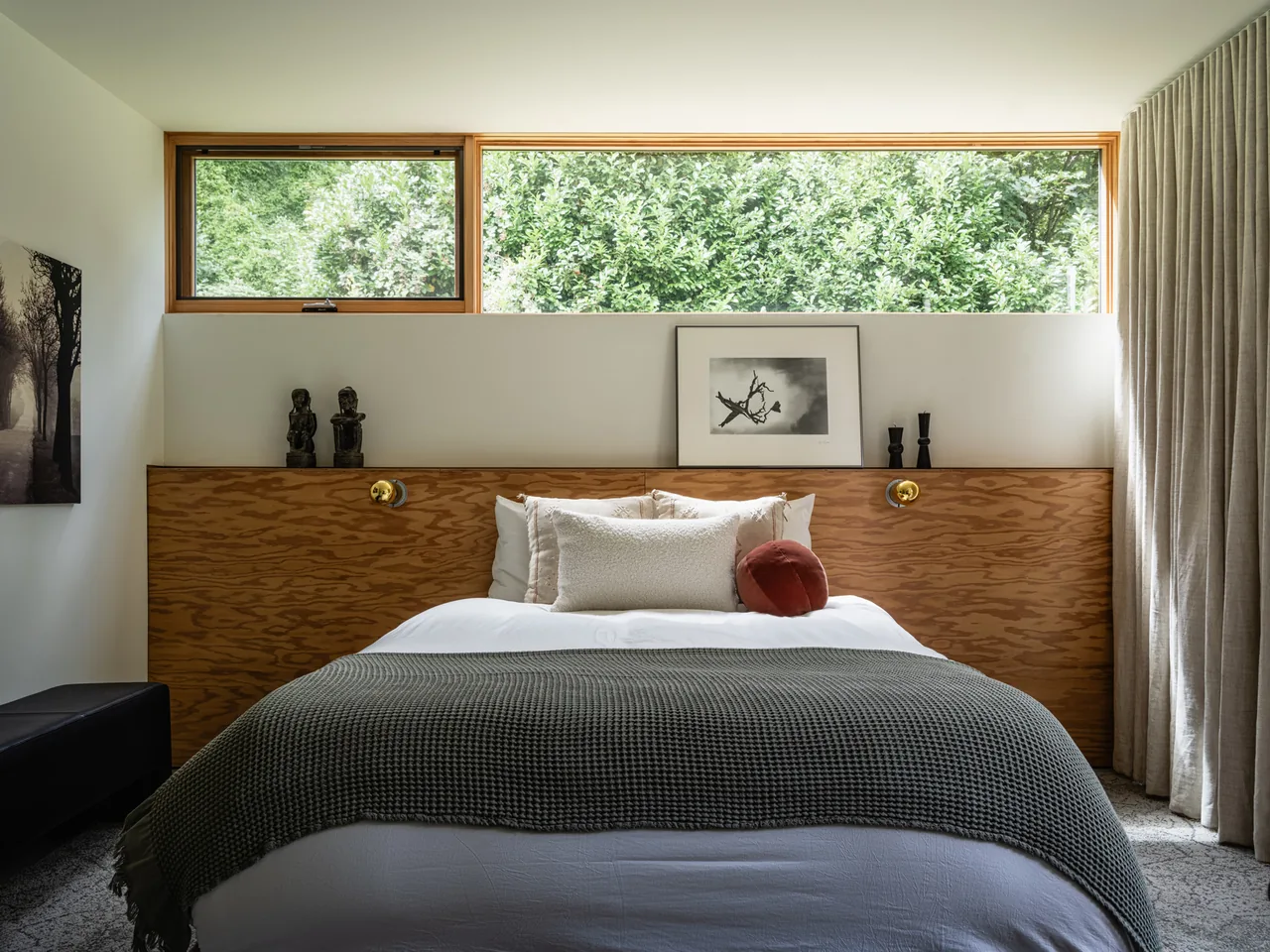 Bedroom at Idea House features a wood-paneled headboard with gold sconces and a wide horizontal window showcasing a garden view.