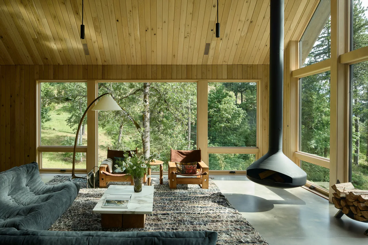 Living room of Sanford Spring project with wood paneling, large glazing, and a suspended fireplace overlooking a forest.