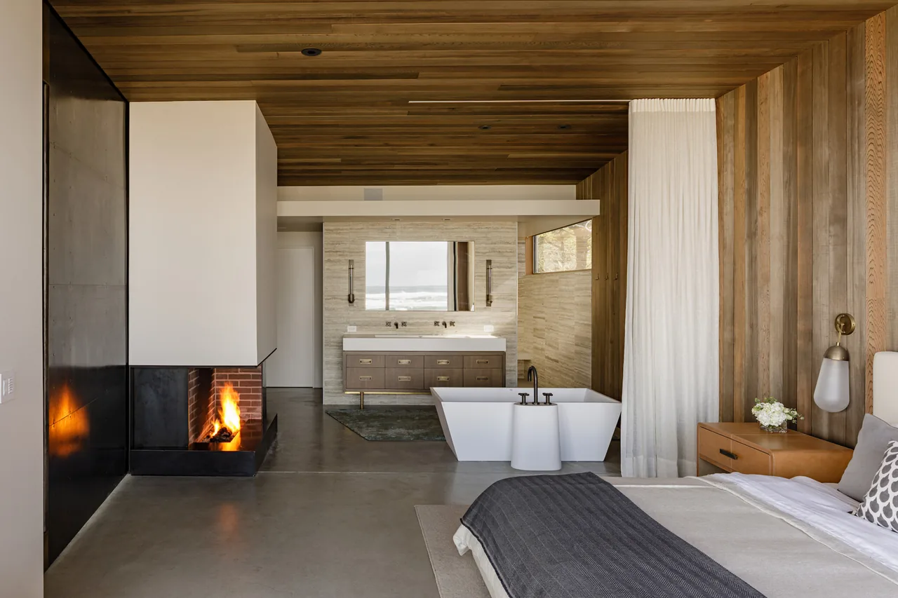 Shelter House master bedroom features a freestanding tub, concrete floor, and a fireplace with wood-paneled ceiling by JHL Design.