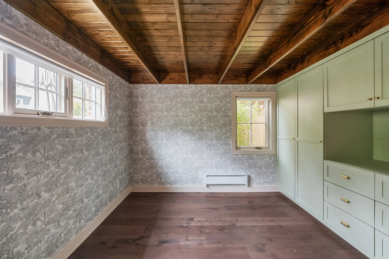 Empty room with dark wood flooring, wallpaper, exposed wooden beam ceiling, and pastel green built-in cabinetry.