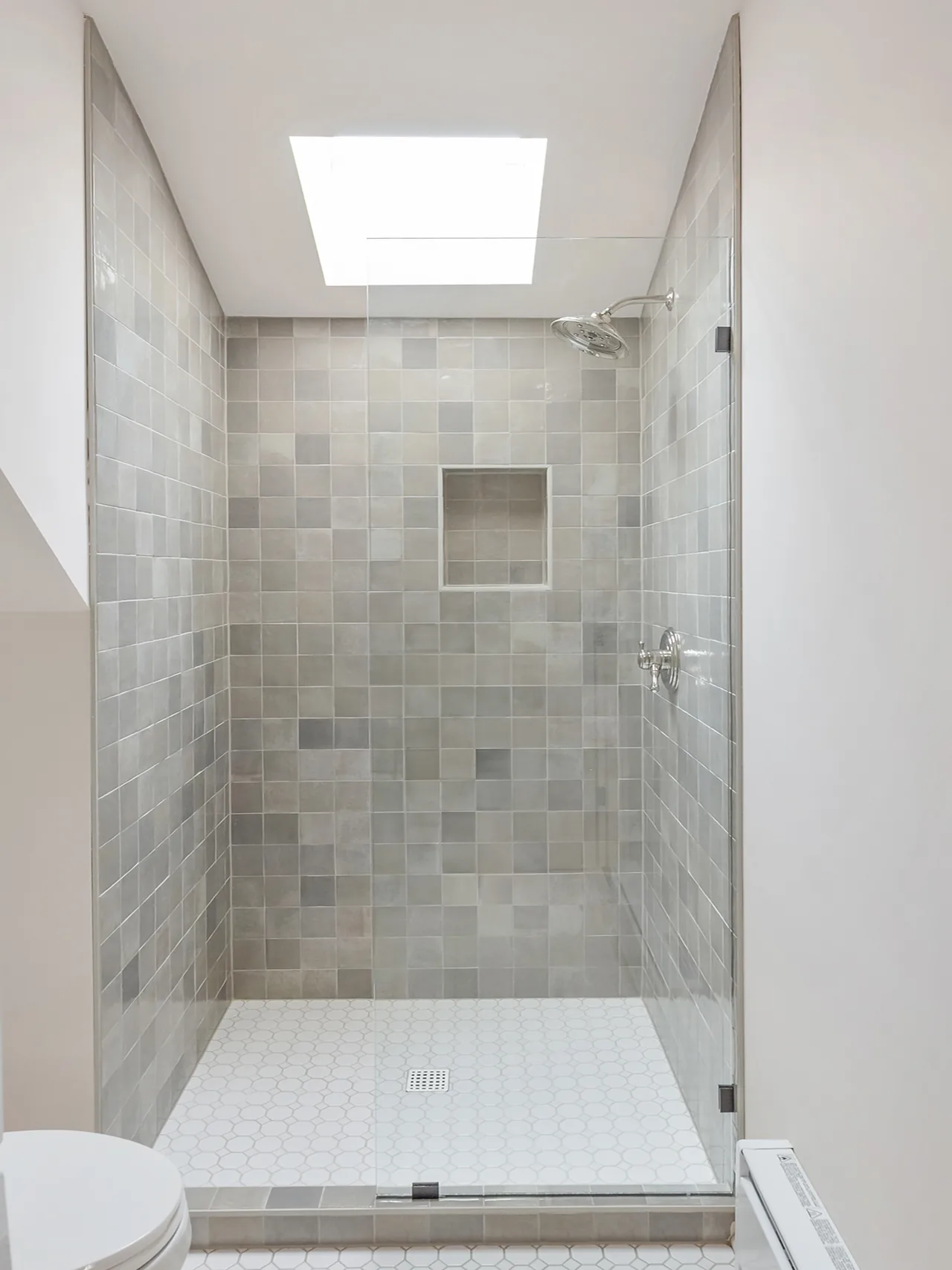 The shower features gray tiled walls, white hexagonal floor tiles, a skylight, and a glass enclosure.