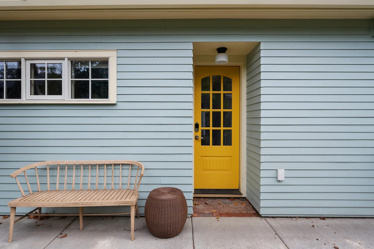 Entry exterior of Buckman ADU with a yellow door, light blue siding, and a wooden bench on the concrete porch.