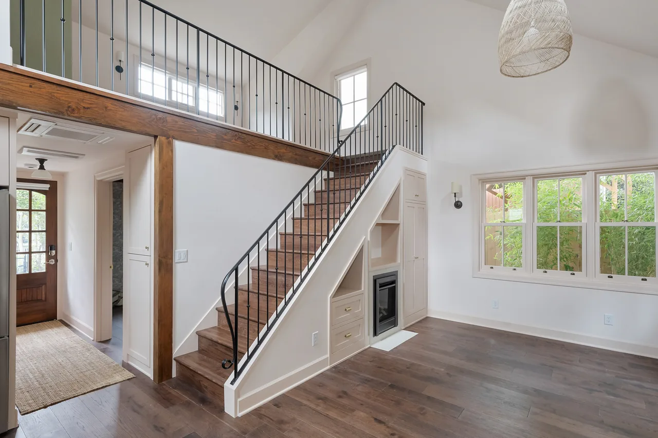 Living room with a two-story ceiling, wooden staircase with metal railing, built-in shelving, and fireplace under stairs.