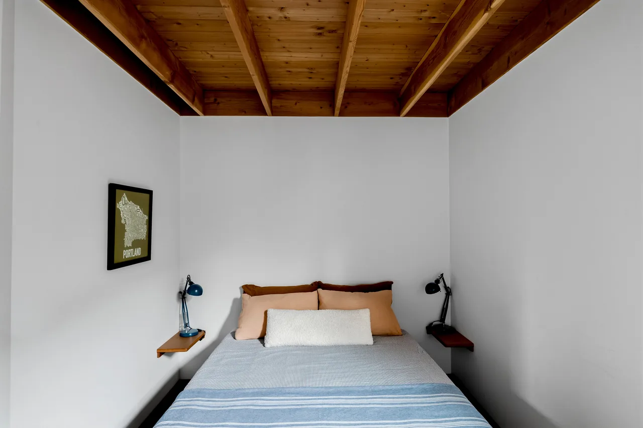 Bedroom with exposed wood beam ceiling, small floating shelves, and two adjustable lamps next to the bed.