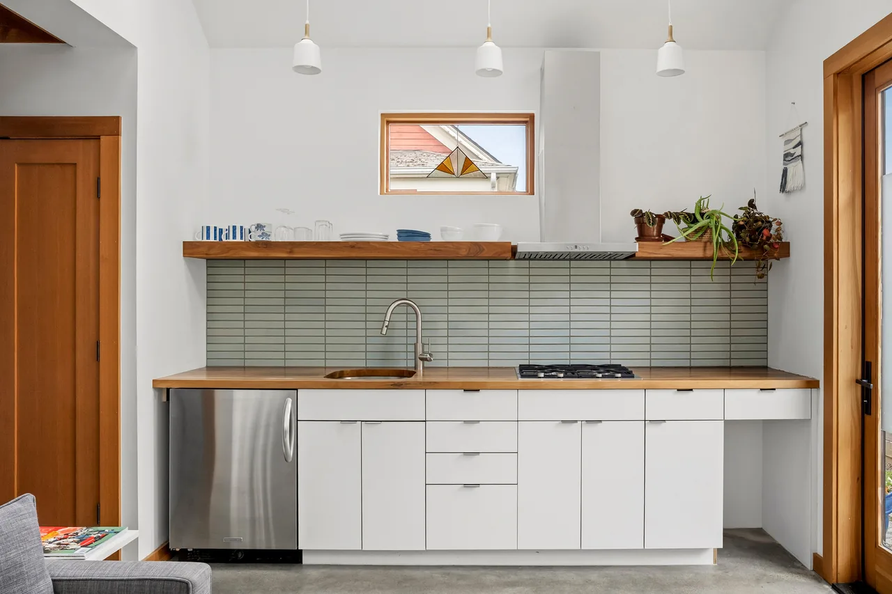 The Irving Park ADU kitchen features white cabinets, wood countertops, and a tiled backsplash with black window frames.
