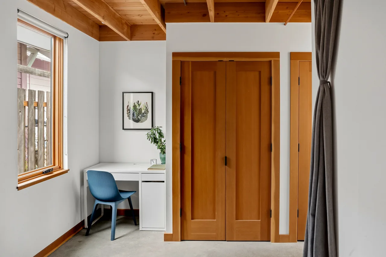 Home office corner showing a white desk, blue chair, wooden closet doors, and exposed ceiling beams with natural wood trim.