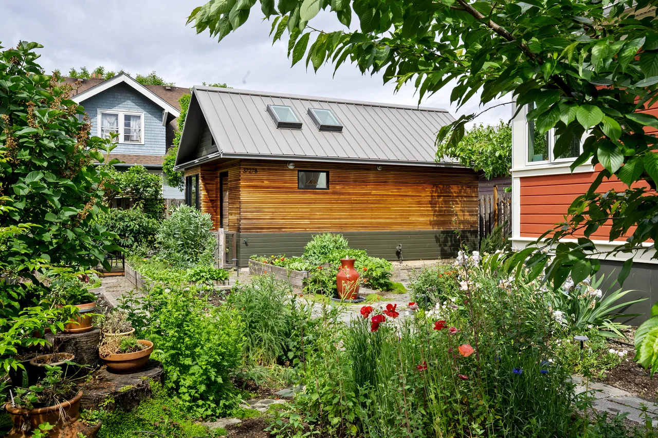 Accessory dwelling unit with wood siding, metal roof, skylights, and raised garden beds with stone pathway in the foreground.