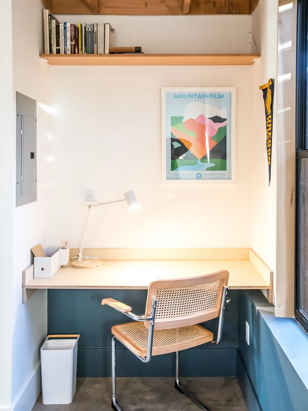 Compact workspace with light wood desk, woven chair, and wall-mounted shelf under exposed wood beams.