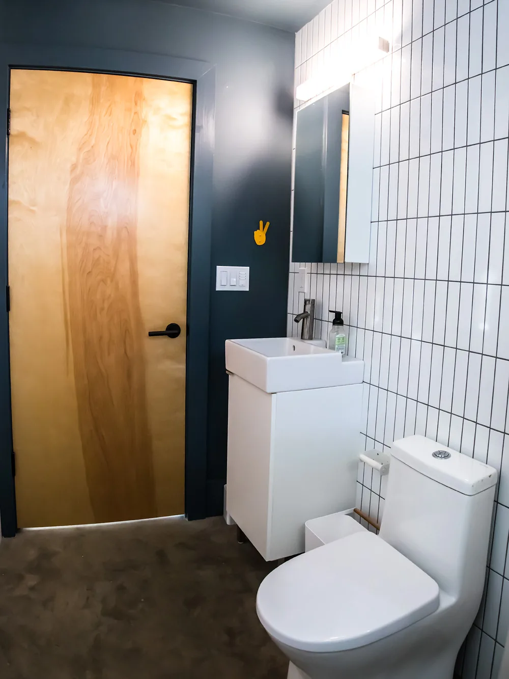 Bathroom with vertical white subway tile backsplash, a wood door, and compact sink vanity next to a modern toilet.