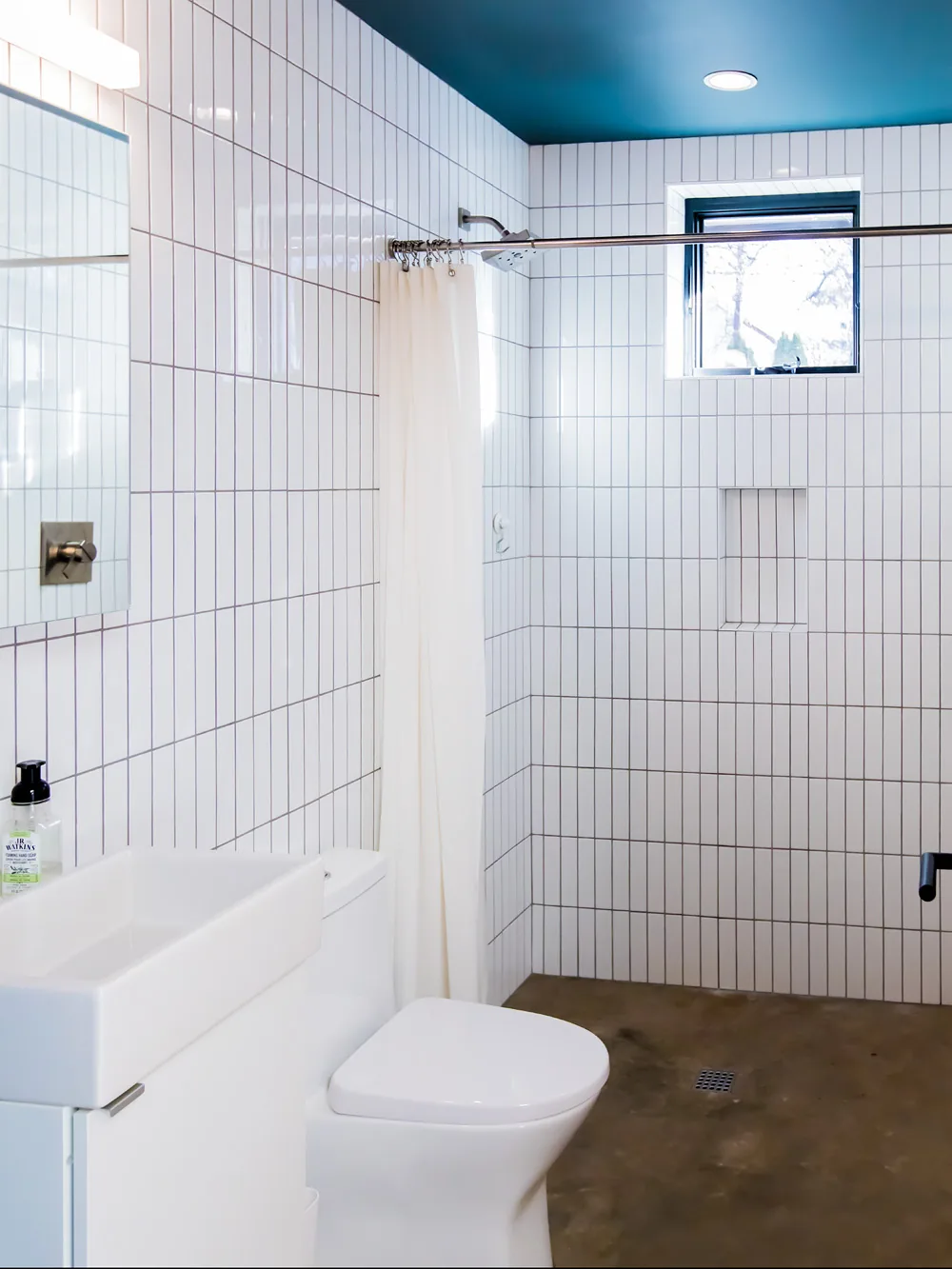 Bathroom features vertical white subway tile on walls, a small vanity sink, and polished concrete floor with a walk-in shower area.