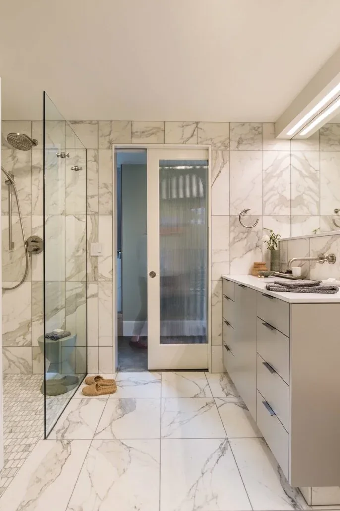 Bathroom with marble tiles, glass shower partition, sliding frosted glass door, and a modern vanity with a narrow sink.