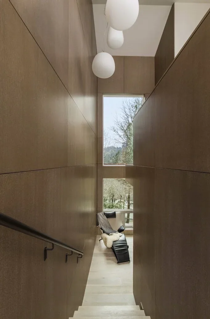 Interior stair with vertical wood paneling and a lounge chair by a large window showing outdoor trees.