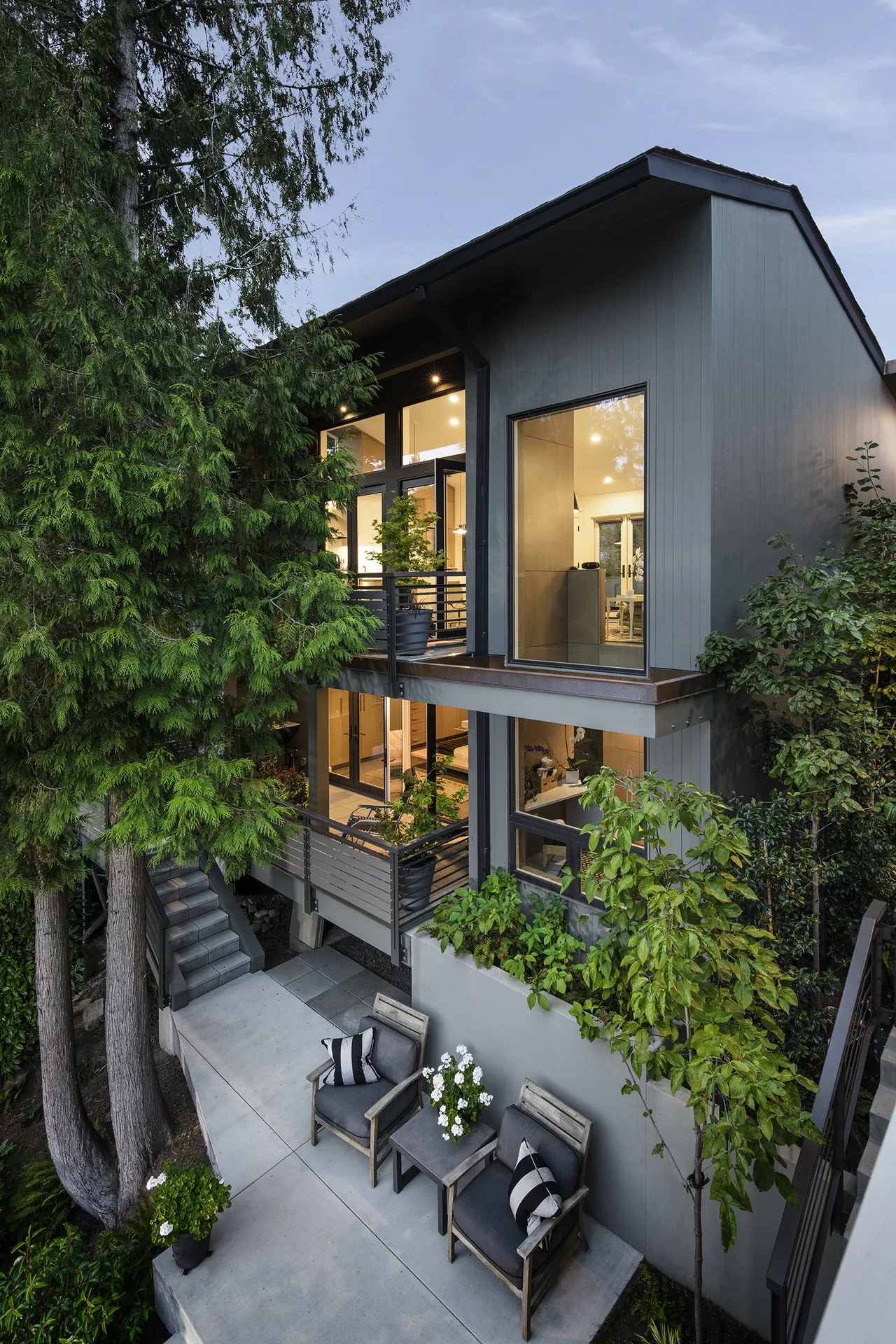 Exterior view of a modern two-story home featuring a balcony and patio seating surrounded by trees and plants.