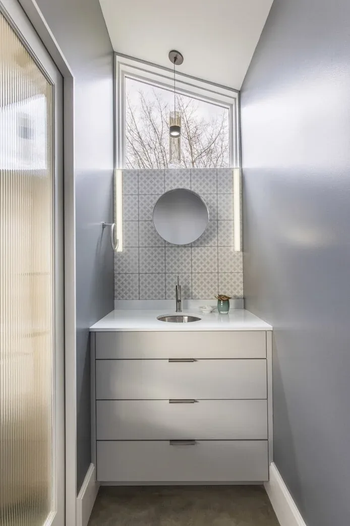 Narrow bathroom vanity with round mirror, pendant light, and patterned tile backsplash under a triangular clerestory window.