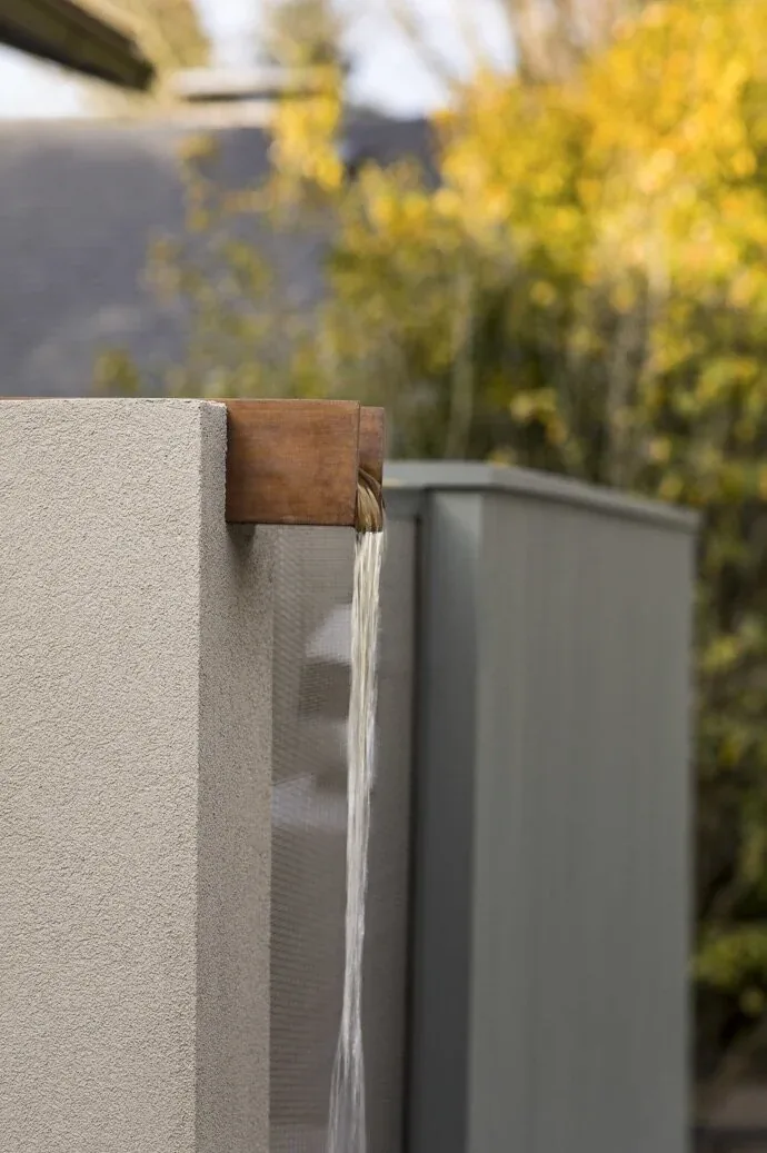 Water streams from a wooden spout embedded in a stucco wall, with foliage in soft focus behind.
