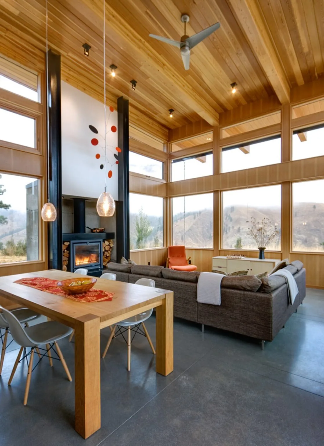 Living and dining space with wood ceiling and large windows showing hillside views, featuring a fireplace and modern pendant lights.