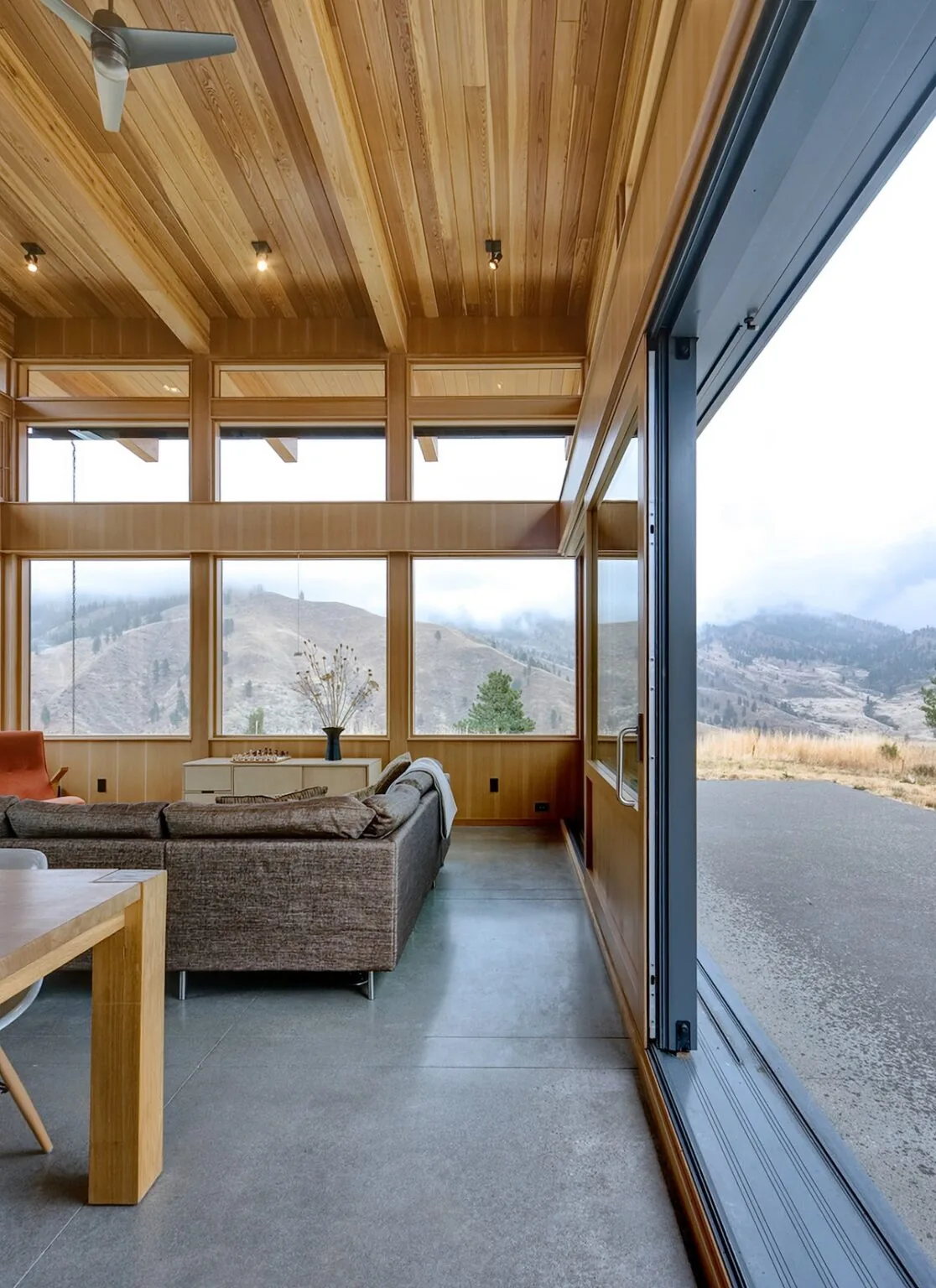 Living room with wood ceiling and window frames, polished concrete floor, and views of a hillside landscape.