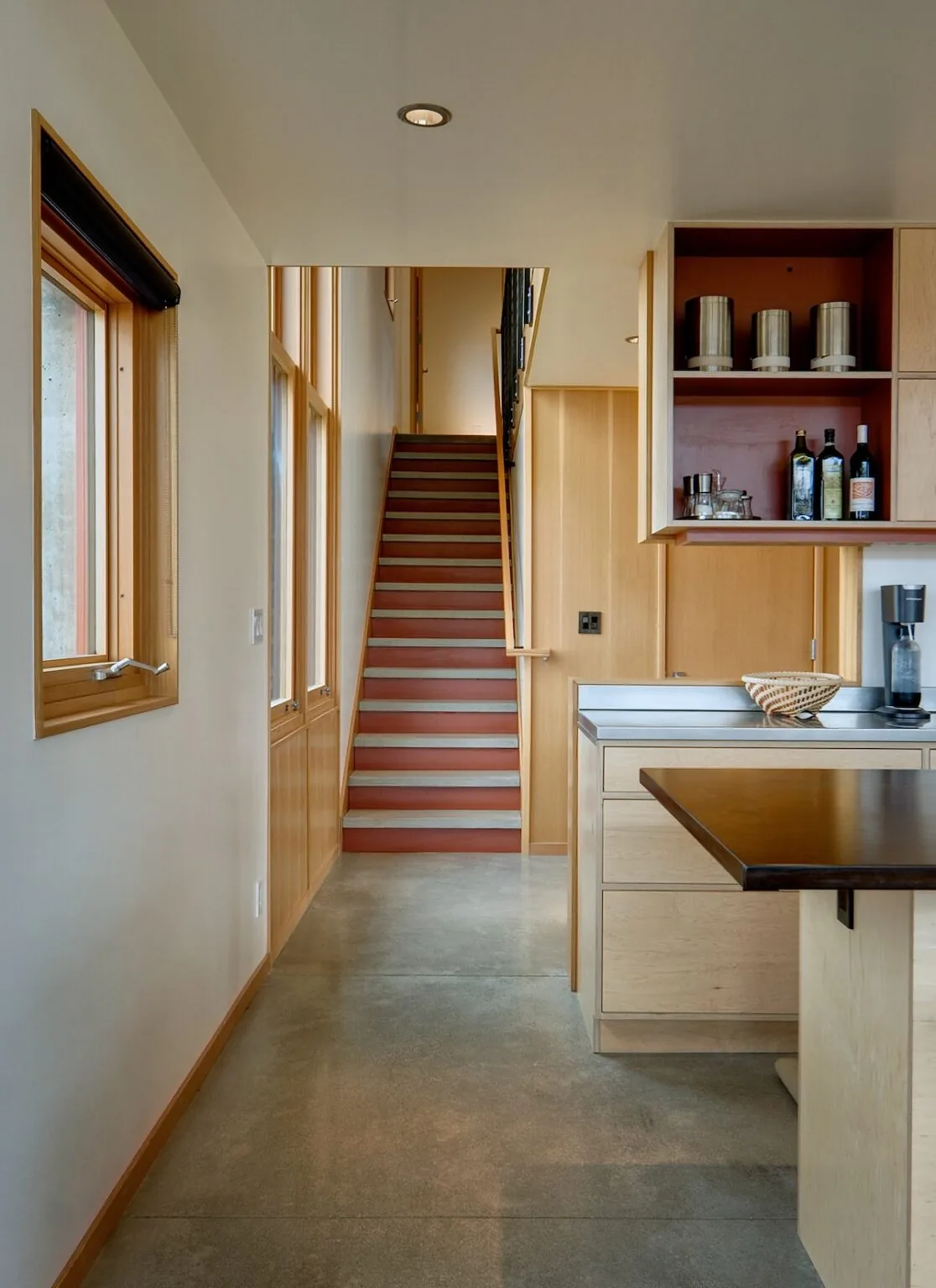 View down a hallway showing a wooden staircase and kitchen cabinetry with stainless steel counter surfaces.